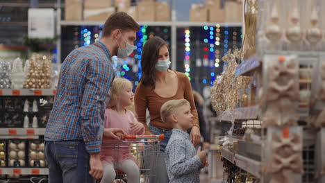 Four-people-mom-dad-and-two-kids-with-a-shopping-cart.-A-happy-family-in-medical-masks-in-the-store-buys-Christmas-decorations-and-gifts-in-slow-motion